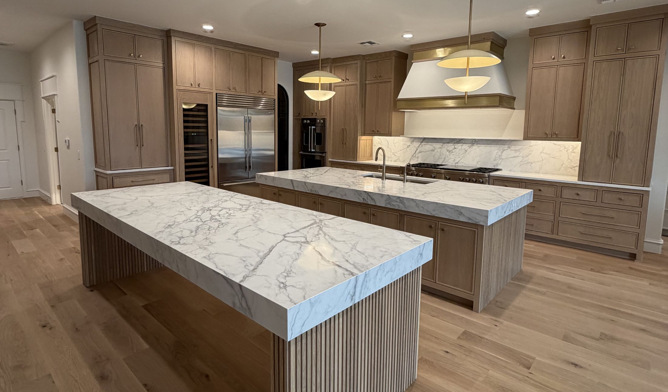 Contemporary rift-oak kitchen with twin marble-topped islands, a fluted island base and a brass-banded range hood, built by Tillison Cabinet Company